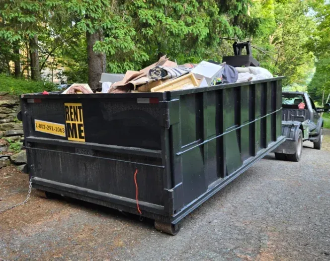 A black dumpster on a trailer, filled with debris, is hitched to a gray pickup truck on a paved driveway.