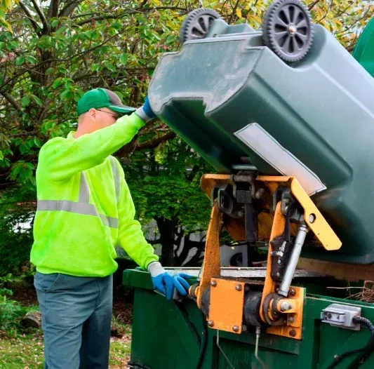 Waste collector in a green vest emptying a dark green trash bin into a garbage truck. Outdoor setting.