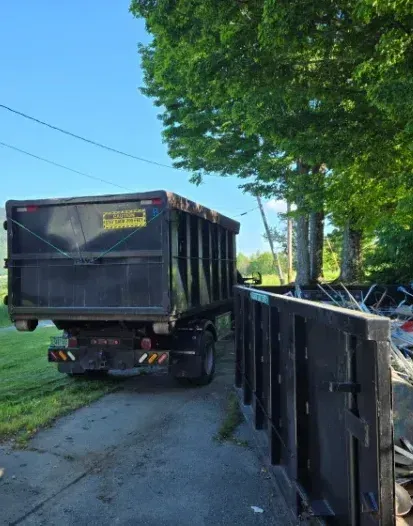 A black dump truck parked on a driveway next to a large pile of debris under a green tree on a sunny day.