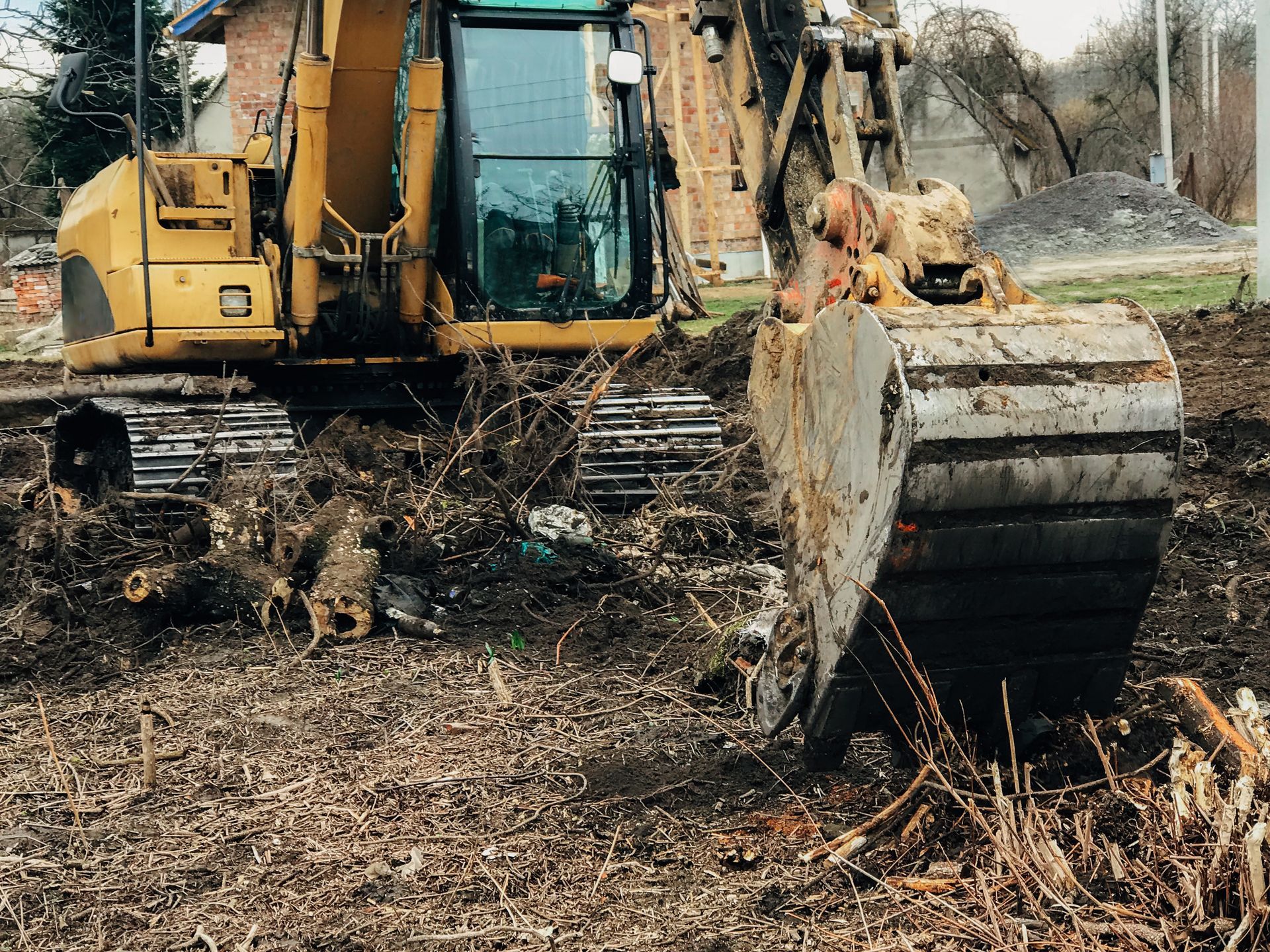 Yellow tracked excavator with a dirty bucket clearing land on a debris-strewn lot