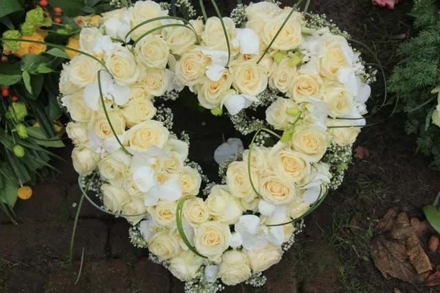 A heart-shaped funeral wreath made of cream-colored roses and white orchids arranged on a dark brick surface.