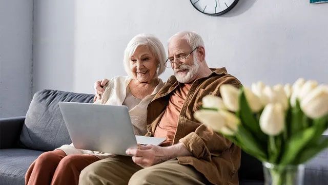 Two people sitting on a sofa and smiling while looking at a laptop computer, with a bouquet of white tulips in the foreground.