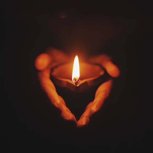 Hands cupped together holding a glowing diya oil lamp against a dark background, forming a heart shape.