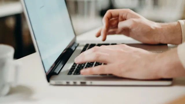 Close-up of hands typing on a laptop keyboard at a desk, with a blurred cup visible in the foreground.