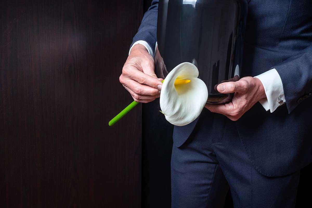 A person in a dark suit holds a black funeral urn and a white calla lily against a dark background.