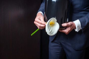 A person in a dark suit holds a black funeral urn and a white calla lily against a dark background.