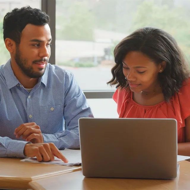 Two people sit at a desk, collaborating on a laptop and reviewing documents in a well-lit office.