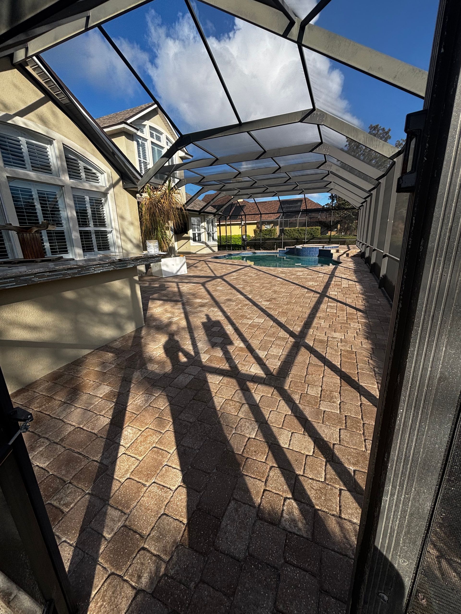 A pool deck with brown pavers enclosed by a screen cage, viewed from an entryway, with a partial view of a house.