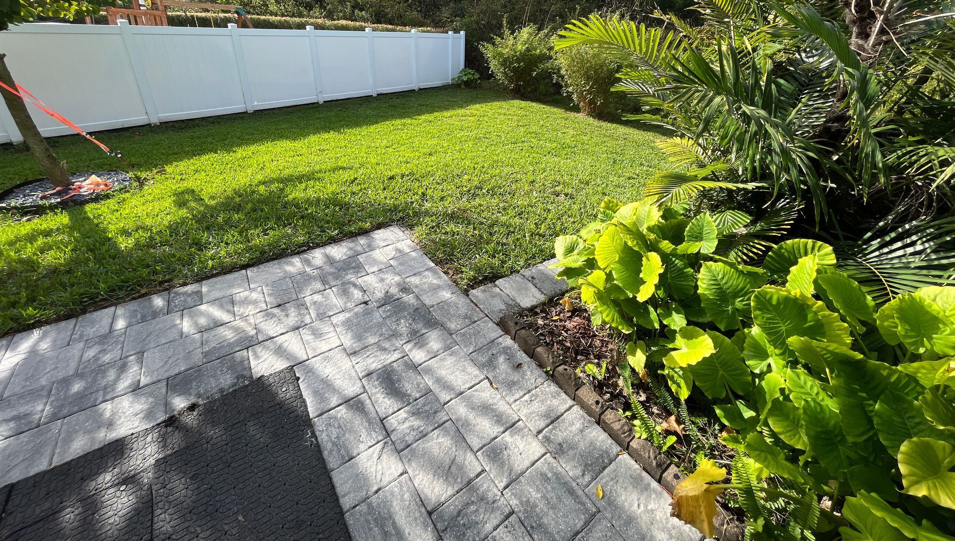 A backyard featuring a paved patio area, lush green grass, vibrant tropical plants, and a white privacy fence.