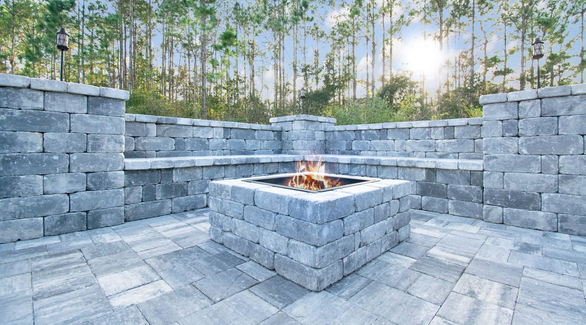 Stone patio with a central fire pit and built-in benches, surrounded by low stone walls and trees at sunset