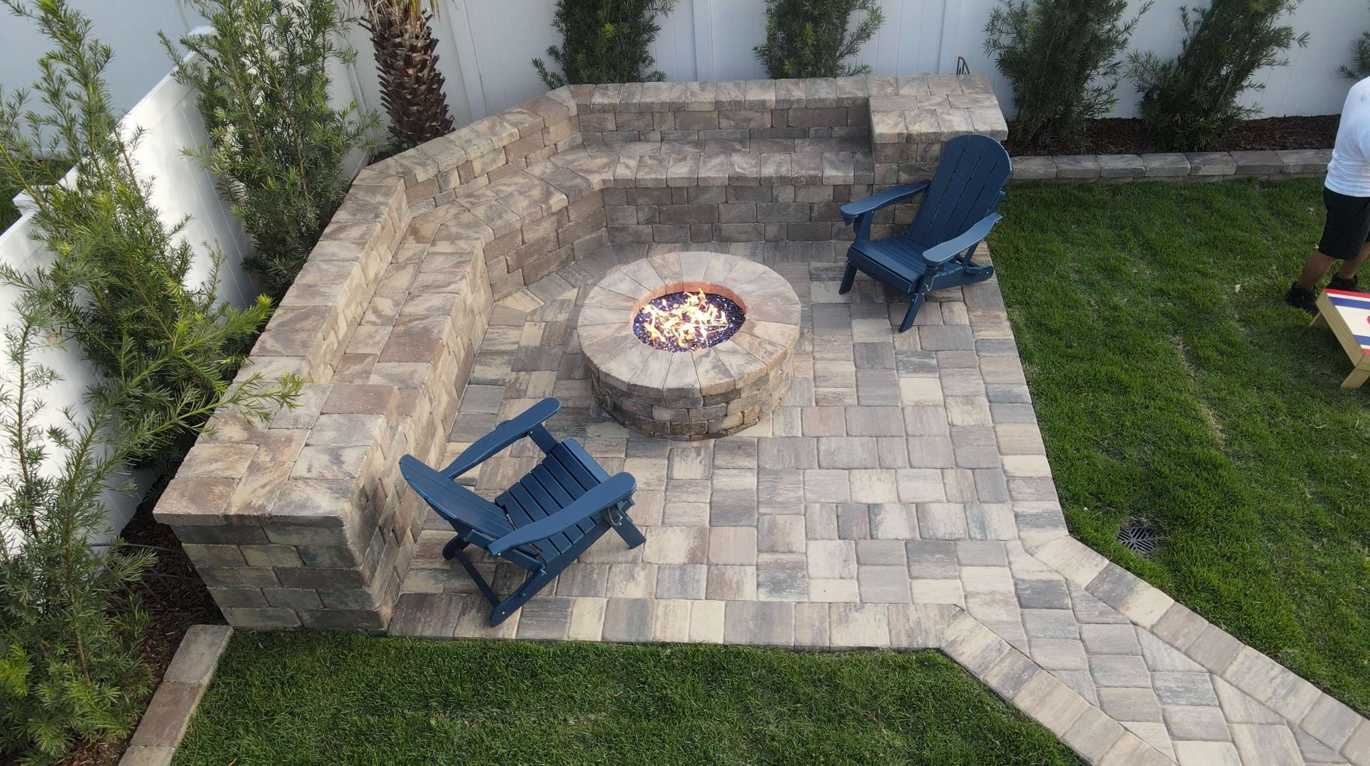 Stone fire pit with built-in seating and blue chairs on a paver patio in a backyard.