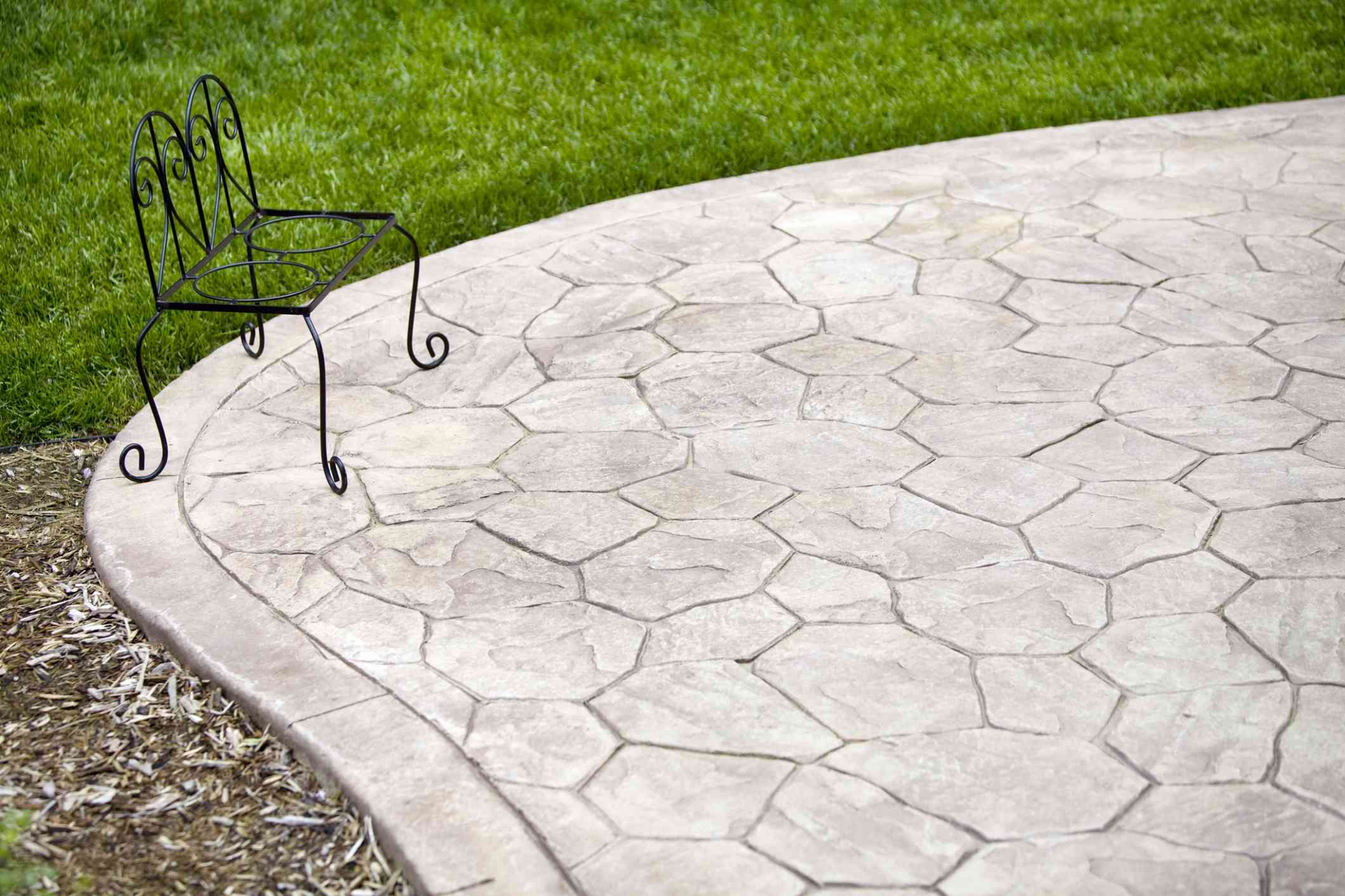 Black wrought-iron chair on a curved stone patio beside green grass.