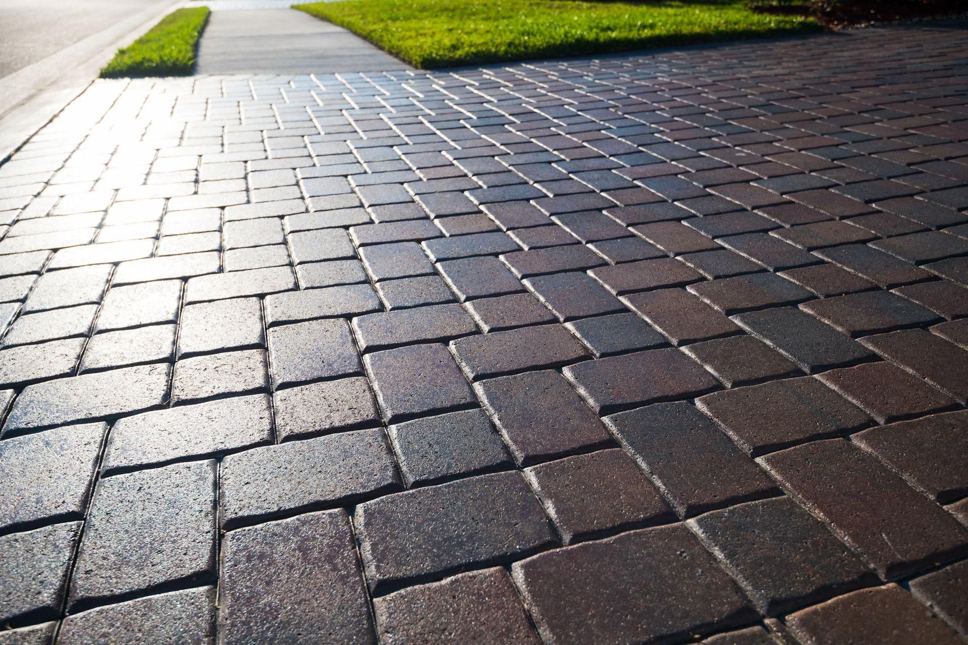 A close-up of a paved driveway made of dark-toned rectangular bricks in a herringbone pattern next to a sidewalk and lawn.