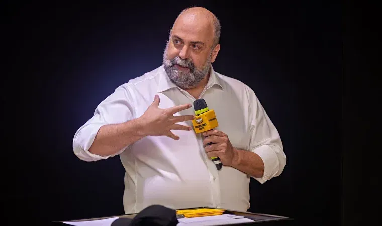 Um homem fala em um microfone amarelo, gesticulando com as mãos. Ele veste uma camisa branca sobre um fundo preto.