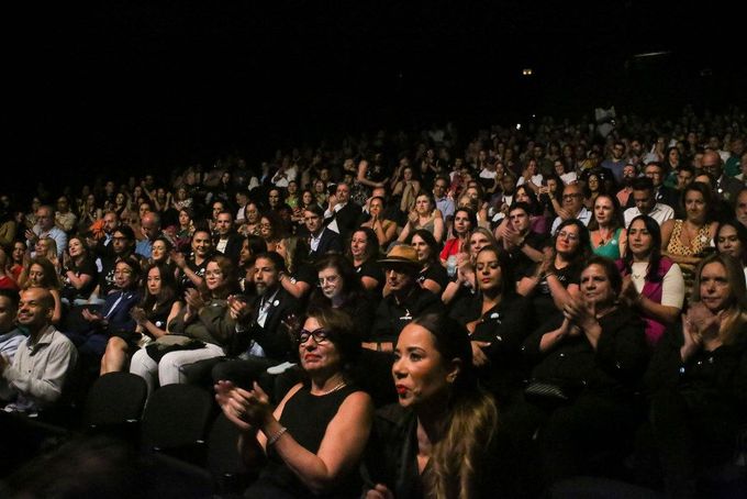 Público aplaudindo em um teatro escuro, muitos vestindo preto. Alguns sorrisos visíveis e tons de pele variados.
