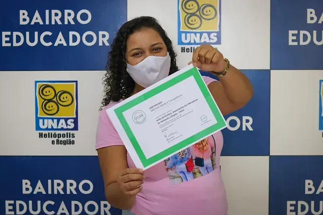 Mulher usando máscara segura um certificado, sorrindo para a câmera, em frente a uma parede azul com a marca "BAIRRO EDUCADOR".