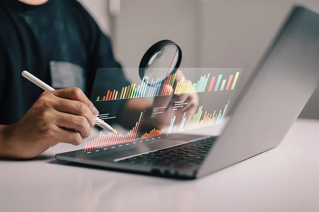 Pessoa usando uma lupa e uma caneta para examinar um gráfico digital em um laptop, com foco em dados financeiros.