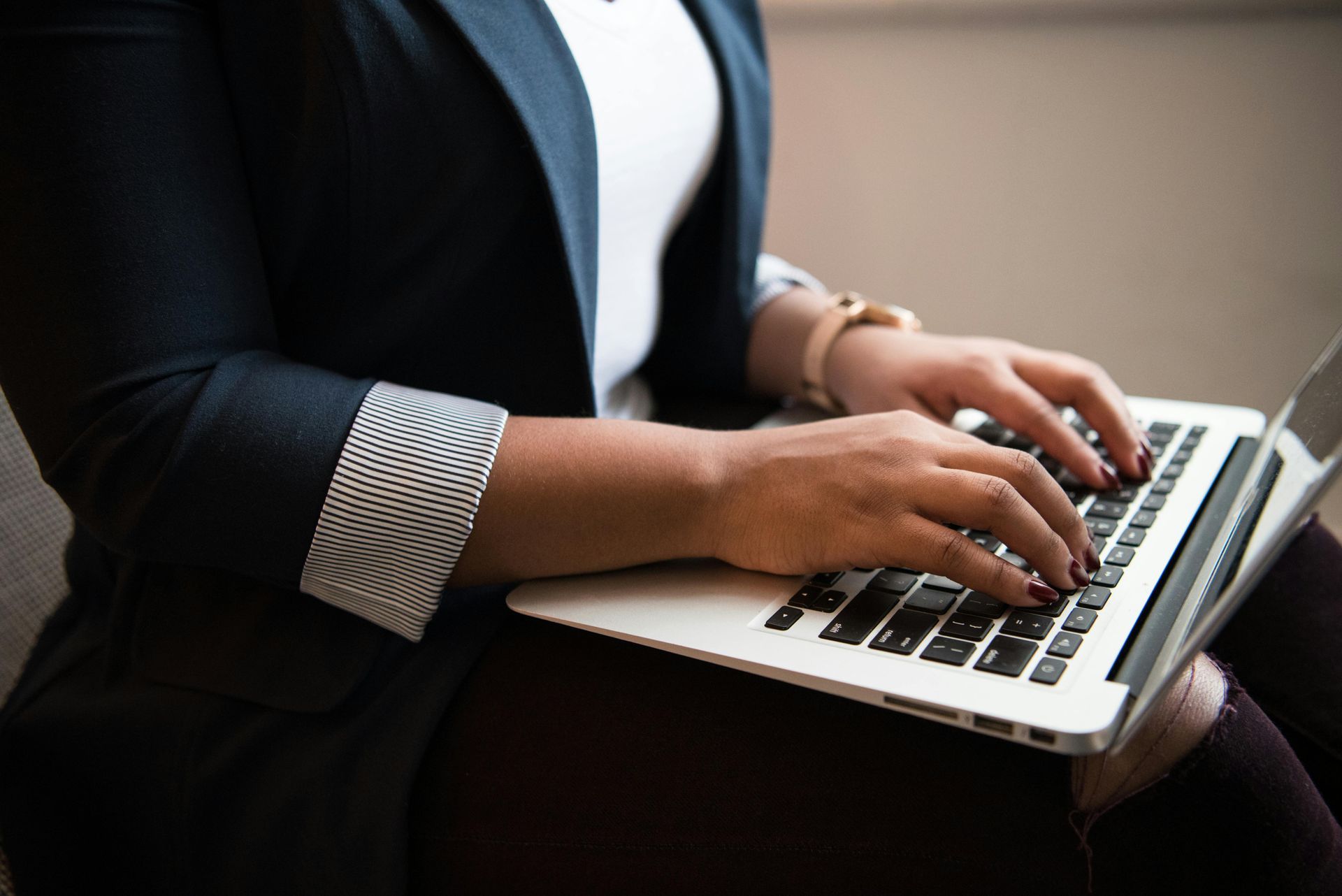Pessoa de blazer digitando em um laptop, em ambiente interno. Foco nas mãos e no teclado.