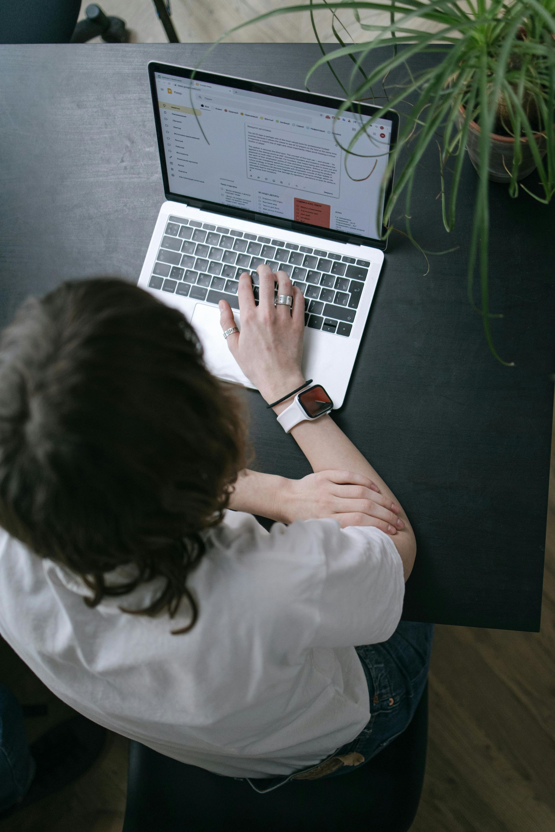 Pessoa trabalhando em um laptop em uma mesa escura, com as mãos no teclado. Há uma planta por perto.