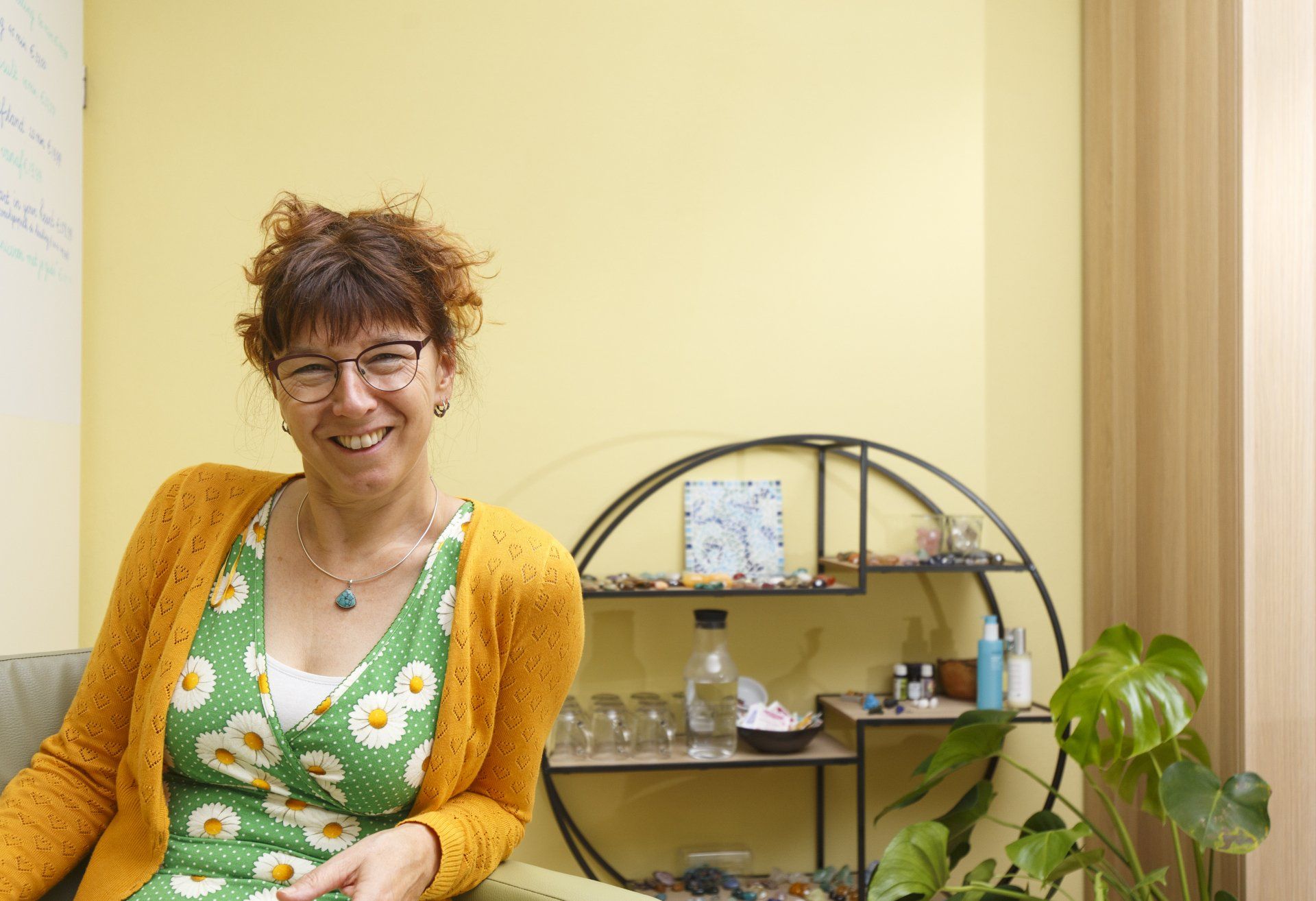 Lachende vrouw, met bril en geel vestje, in een kamer met planken en een plant.