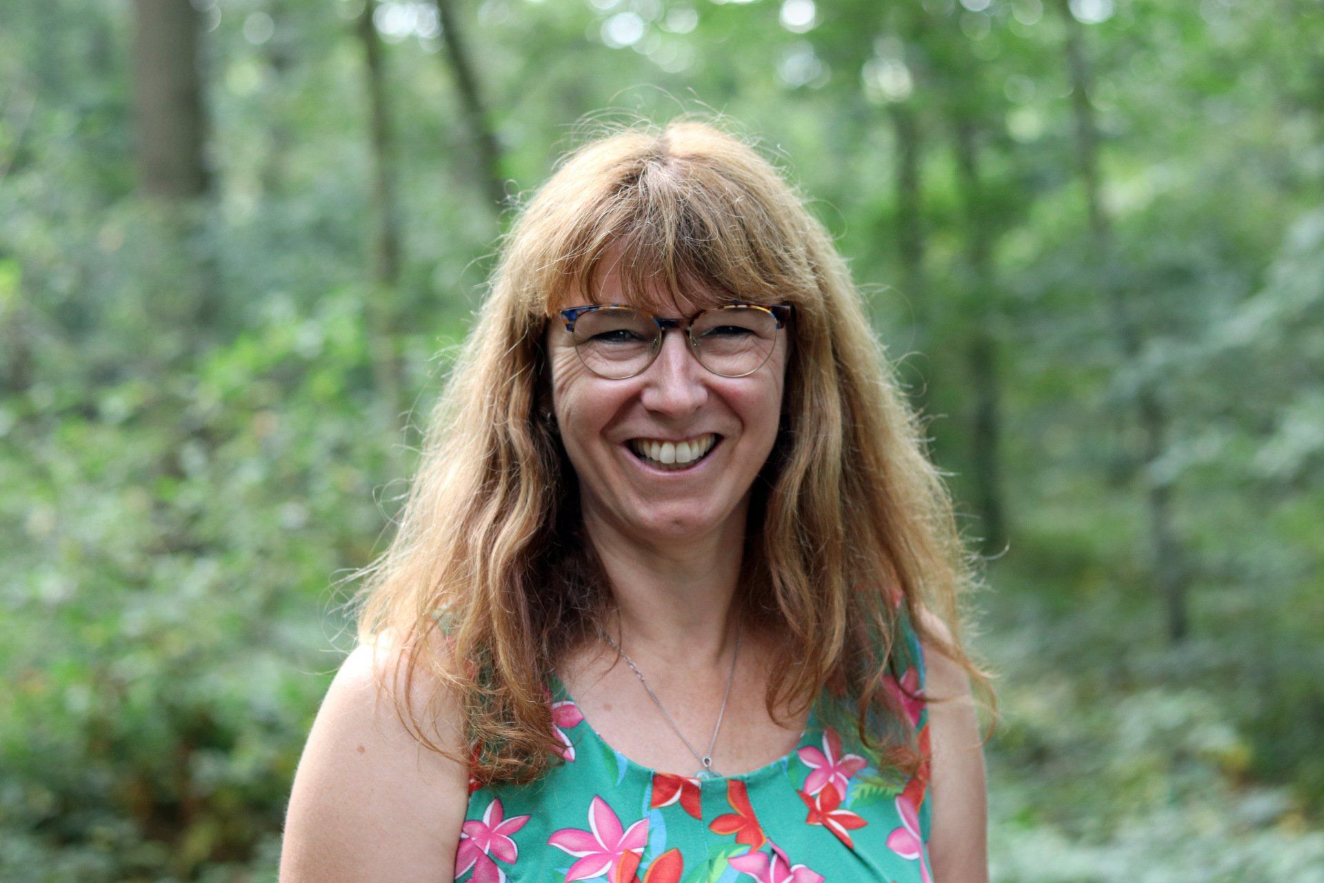 Een vrouw met lang kastanjebruin haar en een bril lacht in een bos; ze draagt ​​een blauwgroene top met bloemenmotief.