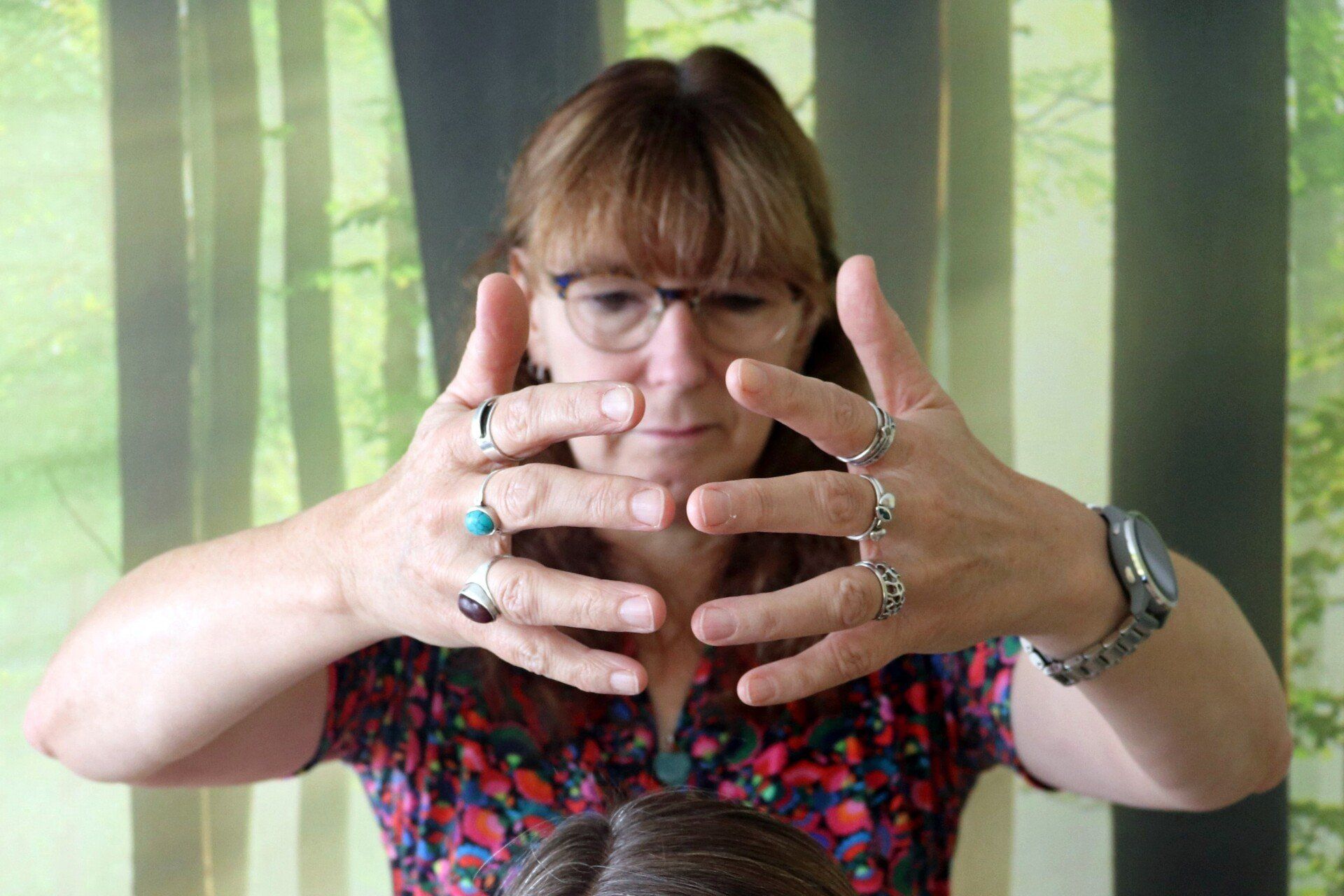 Een vrouw met een bril en ringen strekt haar handen uit naar de toeschouwer, tegen een onscherpe achtergrond van de natuur.
