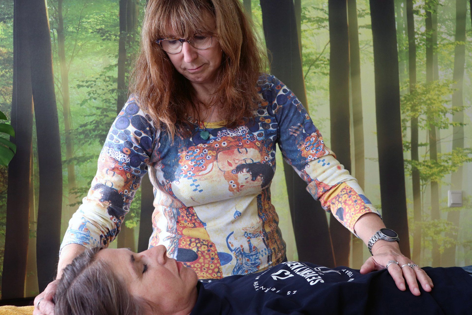 Vrouw voert helende therapie uit op een andere liggende vrouw, voor een muurschildering in het bos.