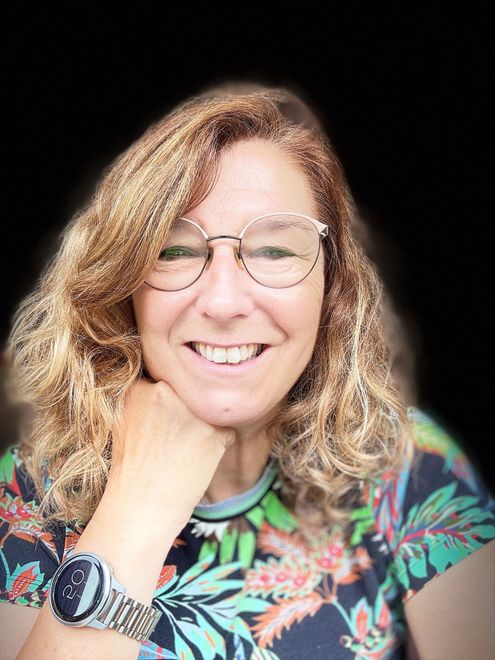 Vrouw met bril glimlacht, hand op haar hoofd, draagt een blauwe trui met bloemenkraag.