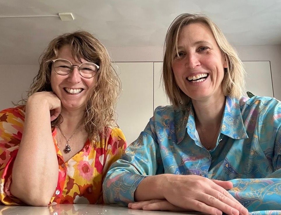 Twee lachende vrouwen, de een met een bril en een bloemetjesblouse, de ander in een blauw bloesje.