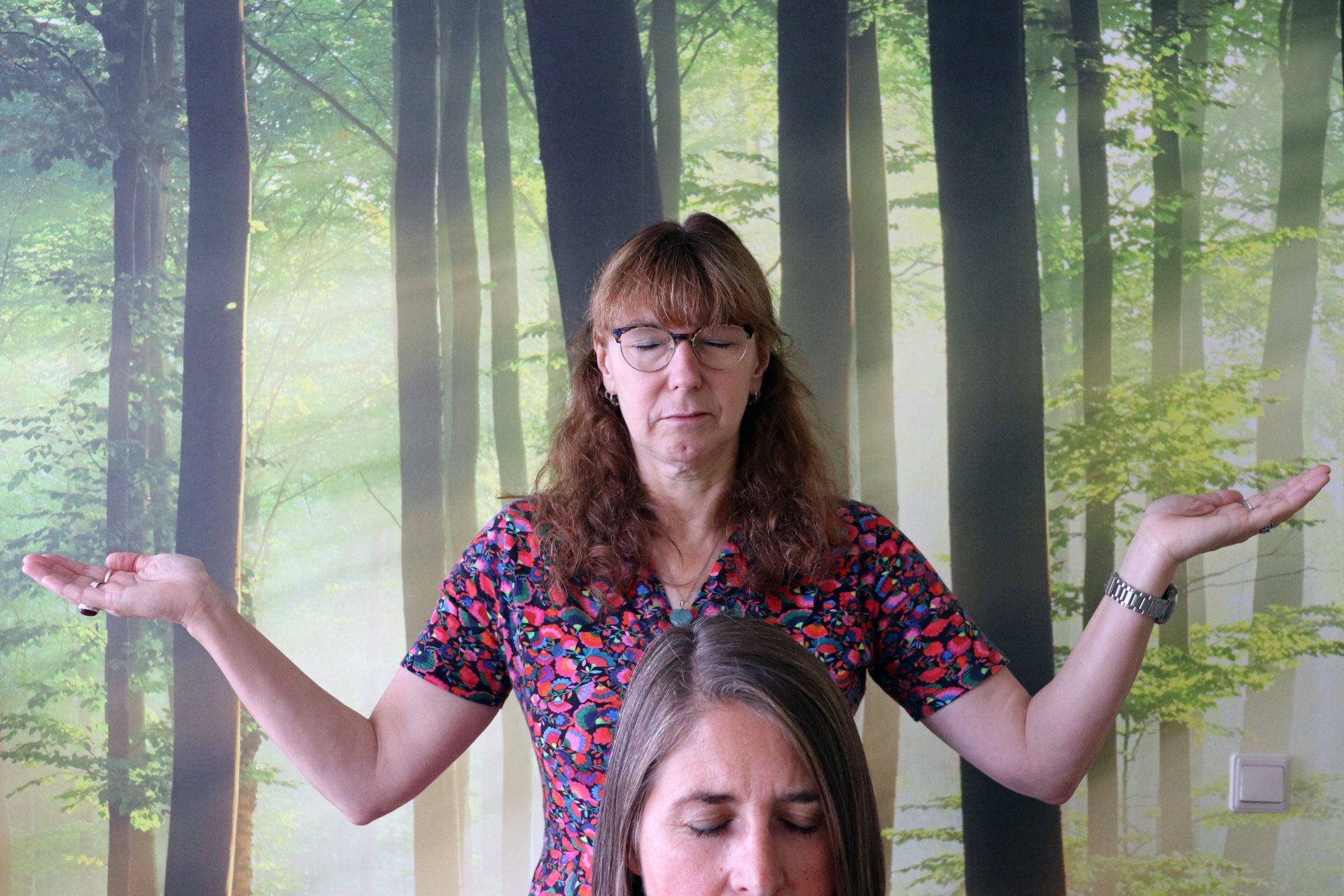 Vrouw met uitgestrekte armen boven een andere vrouw, beide ogen gesloten, voor een muurschildering in het bos.