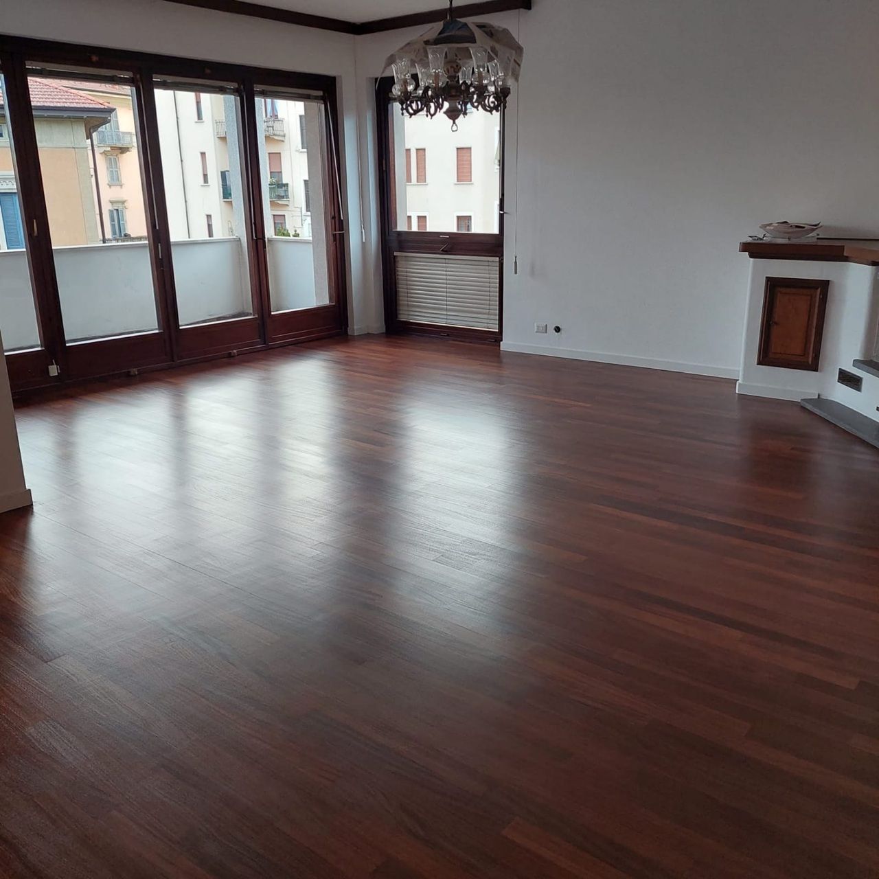 Soggiorno vuoto con pavimento in legno lucido, ampie finestre e lampadario.