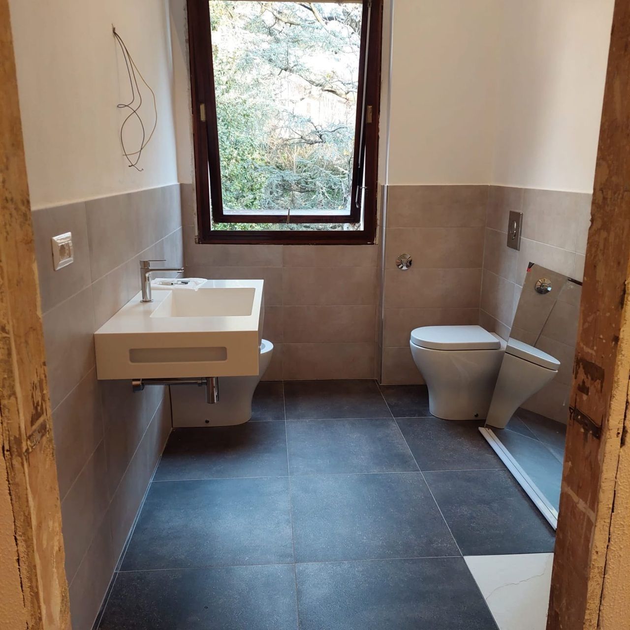 Piccolo bagno con lavabo, WC, doccia e finestra, rivestito con piastrelle beige e grigie.