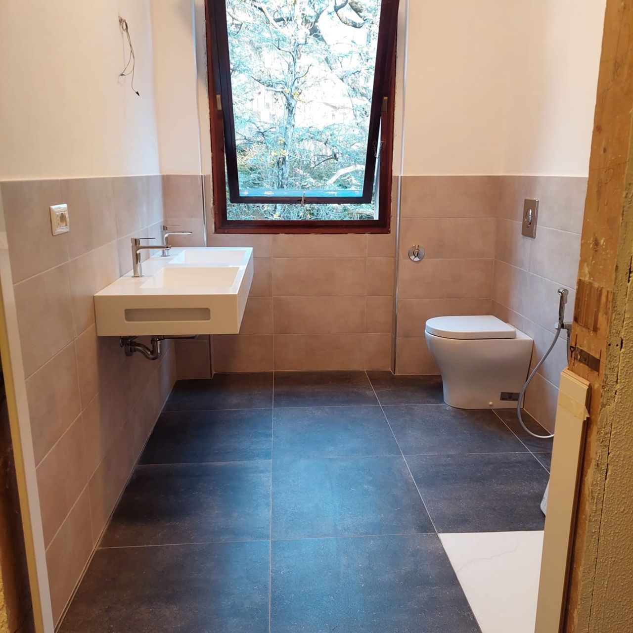 Bagno moderno vuoto con lavabo, WC, ampia finestra e pareti rivestite con piastrelle beige.