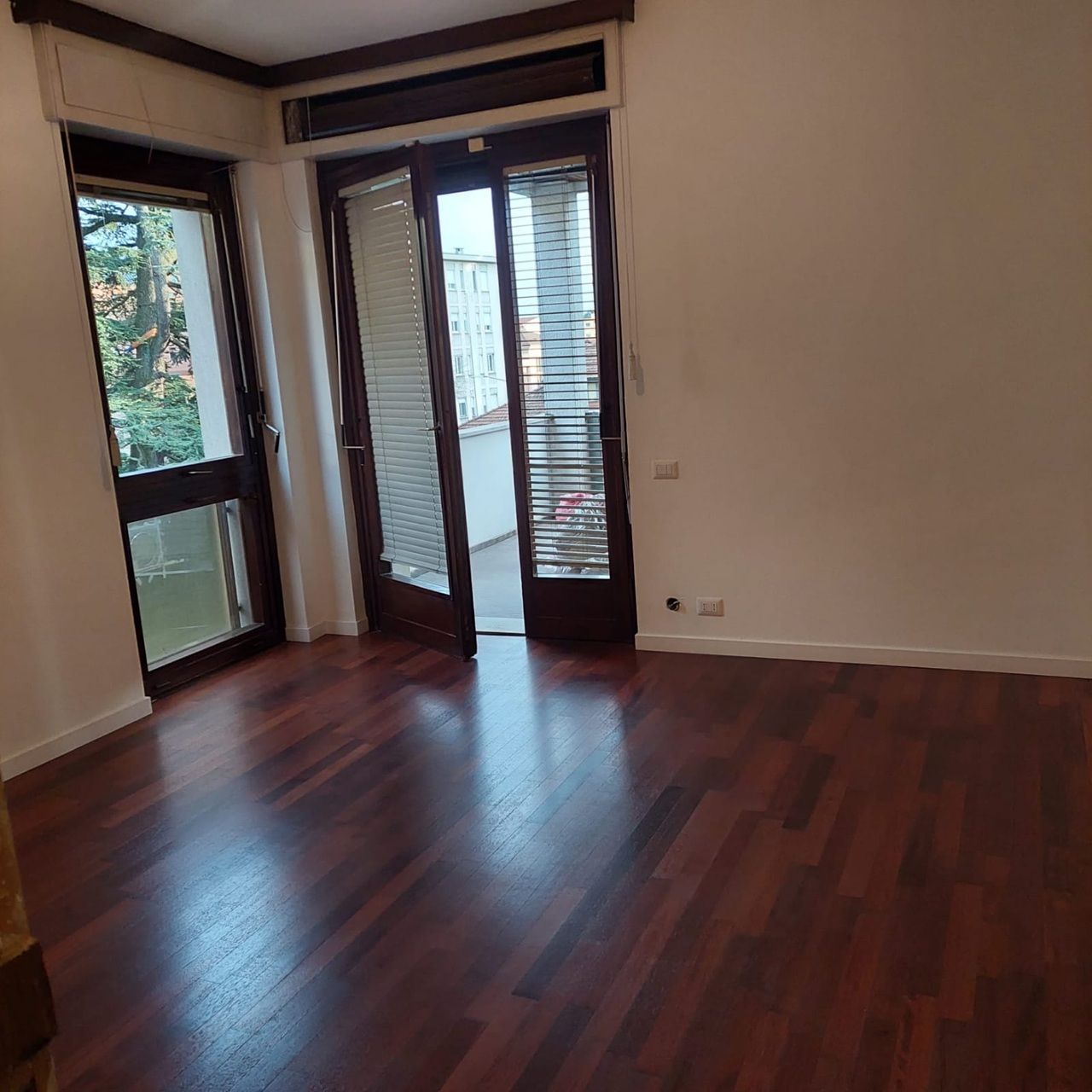Stanza vuota con pavimento in legno scuro lucido, pareti beige e ampie finestre/porte che si aprono su un balcone.