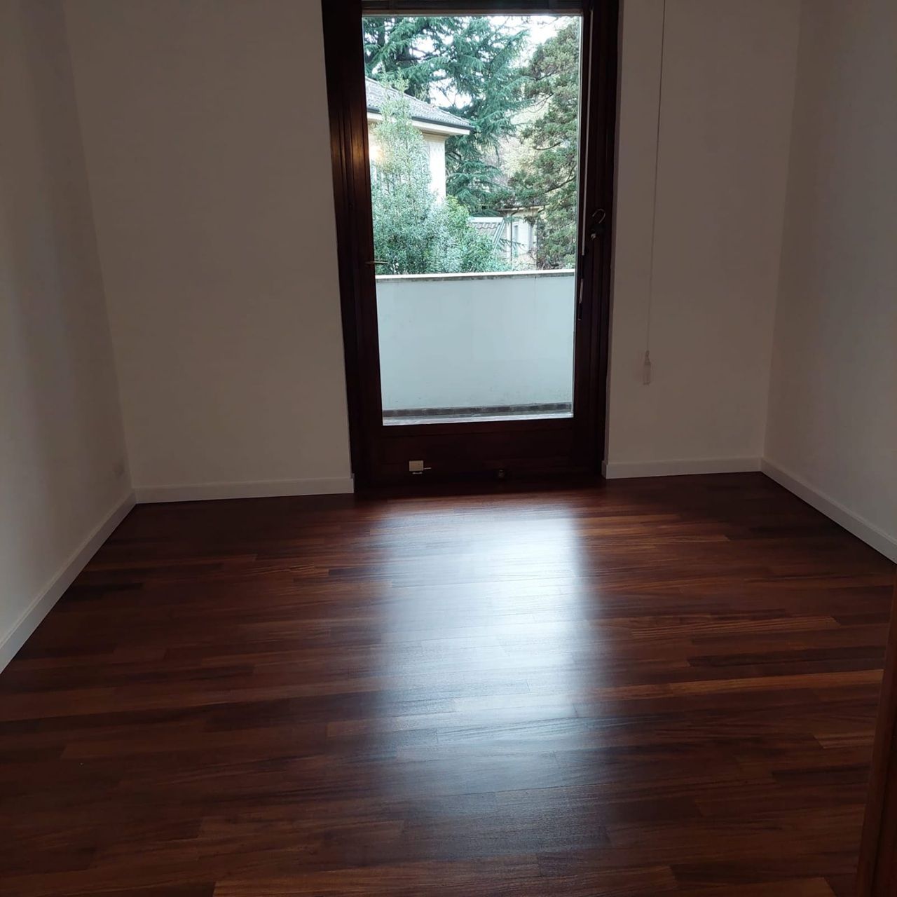 Stanza vuota con pavimento in legno scuro e porta a vetri che si apre su un balcone e sugli alberi.