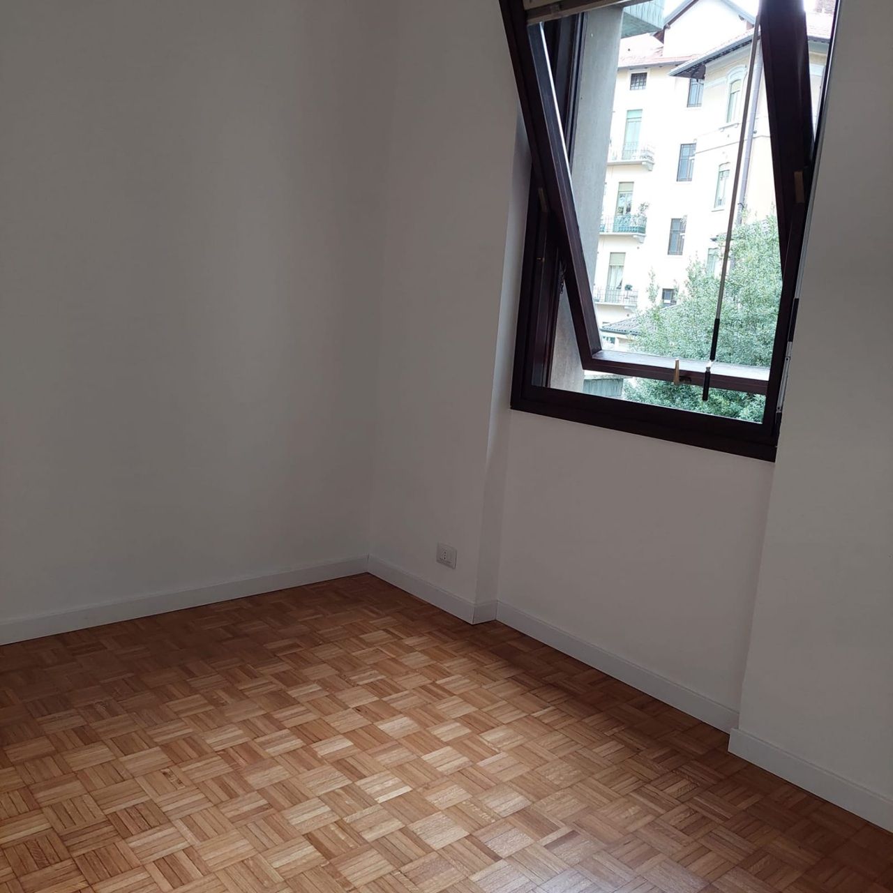 Stanza vuota con pavimento in parquet e finestra aperta che si affaccia sugli edifici circostanti.