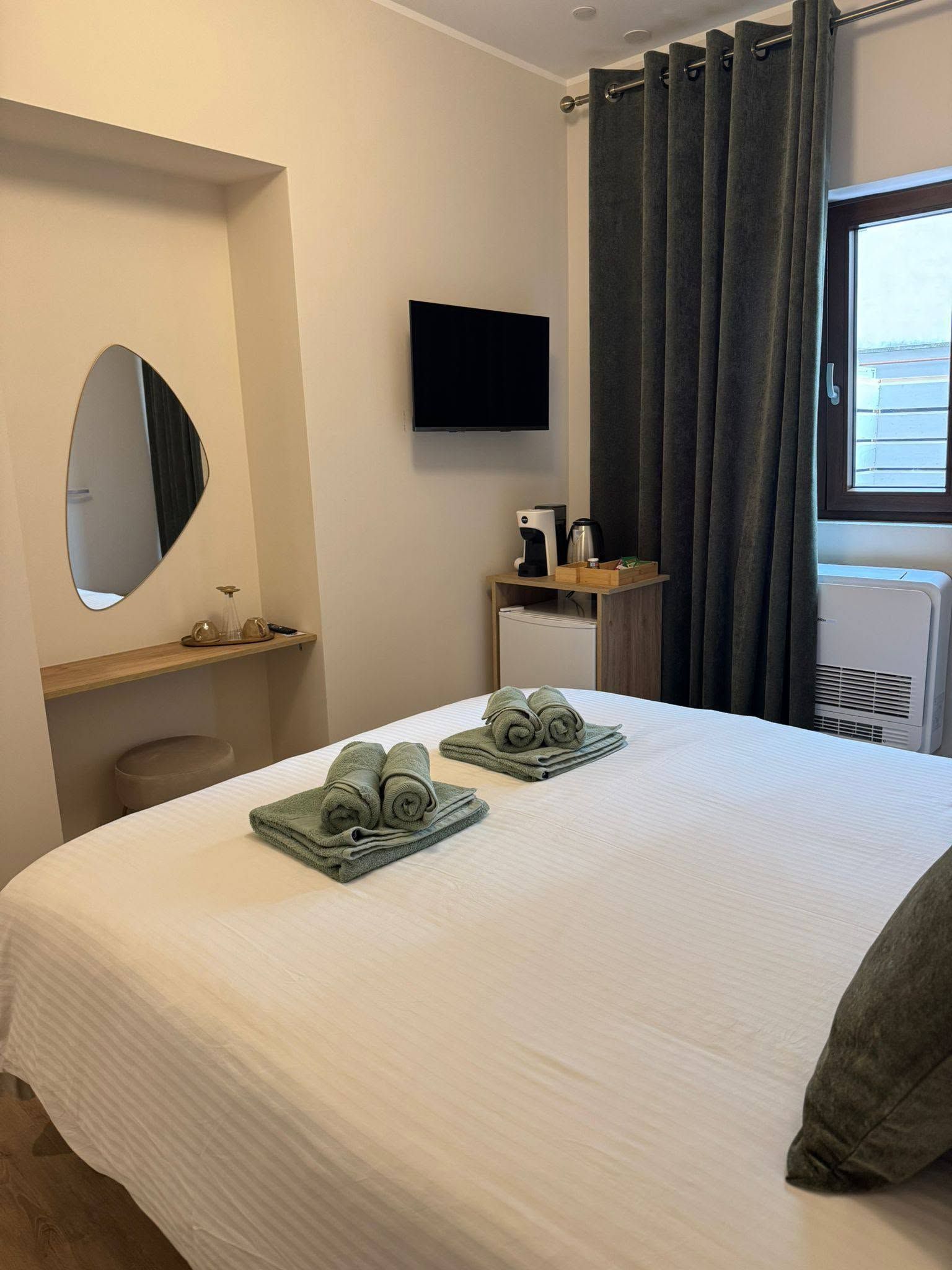 Camera d'albergo ordinata con letto, TV a parete, scrivania, specchio e finestra con tende grigie.