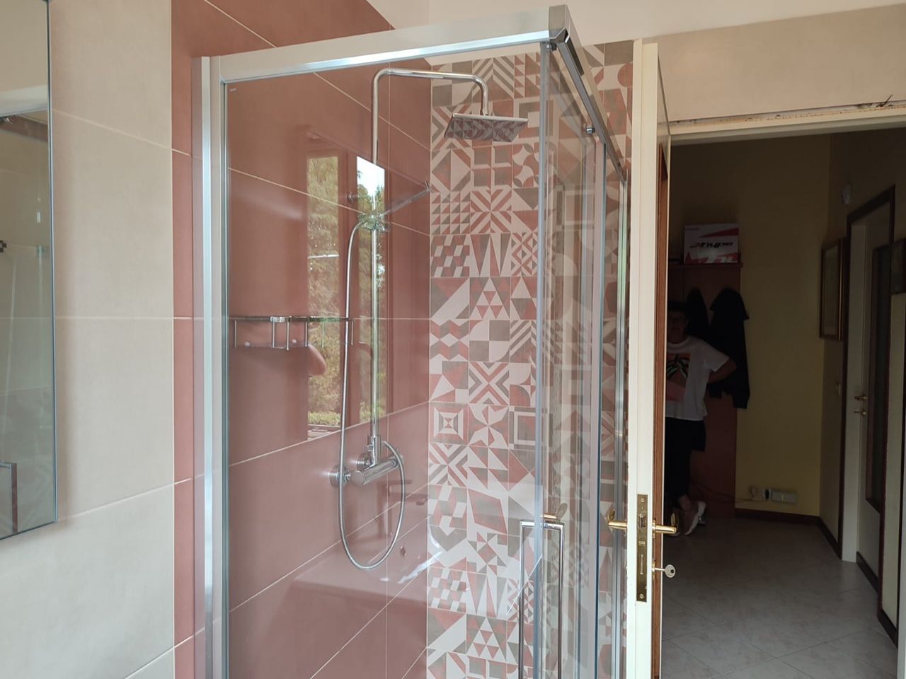 Box doccia in vetro in un bagno piastrellato con pannello divisorio decorato e porta aperta sul corridoio.