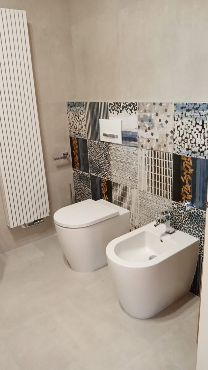 Bagno moderno con WC e bidet bianchi, pavimento beige e parete decorativa rivestita con piastrelle a mosaico colorate.
