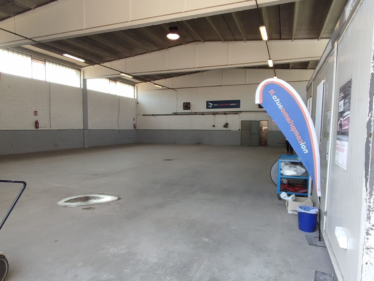 Capannone vuoto all'interno di un magazzino con pavimento in cemento, pareti bianche e uno striscione promozionale blu vicino alla parete di destra.