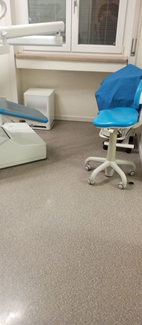 Sala visite mediche con finestra, termosifone bianco, sgabello rivestito di blu e attrezzature dentistiche/mediche.