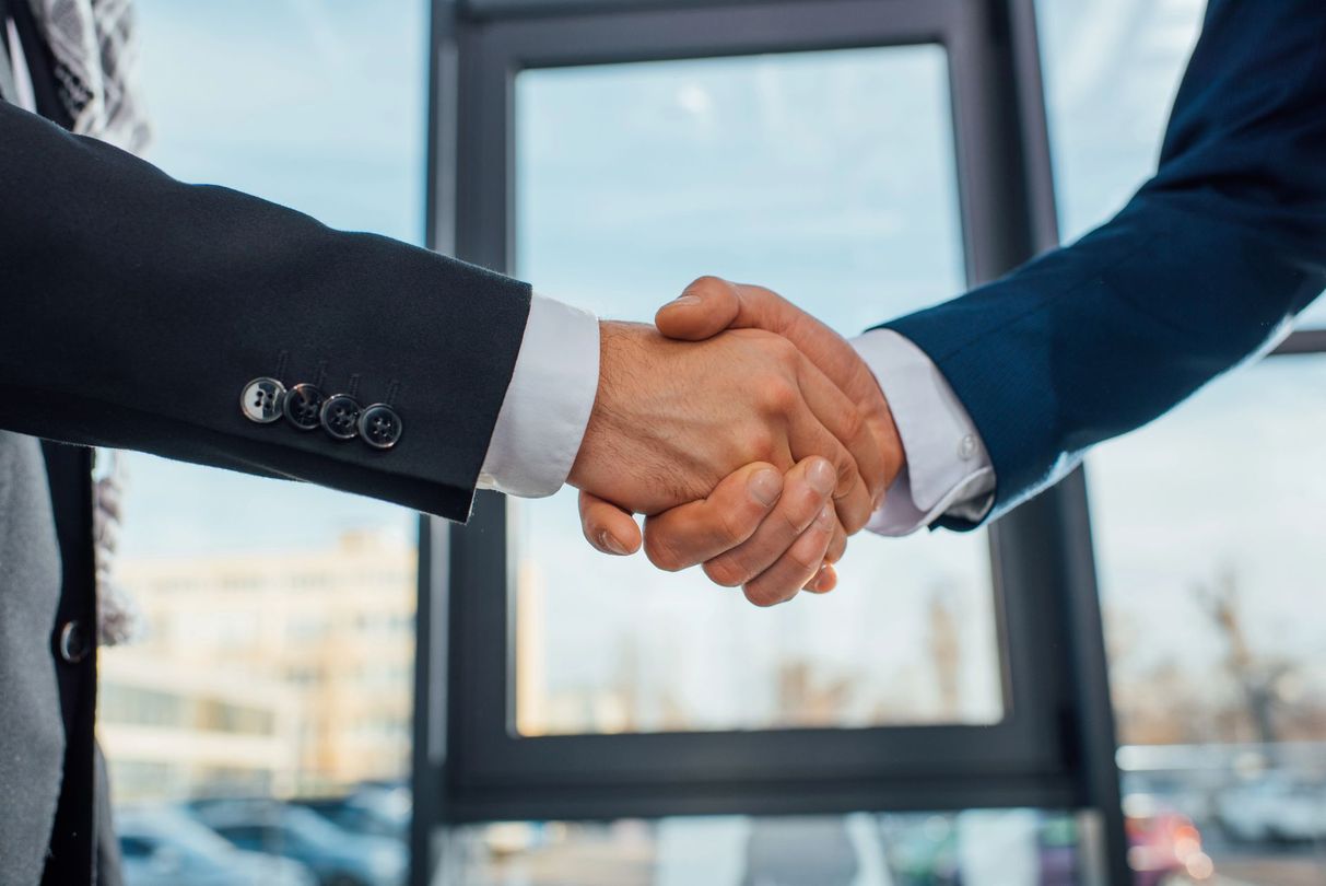 Due uomini d'affari in abito scuro si stringono la mano in un ufficio con una finestra sullo sfondo.