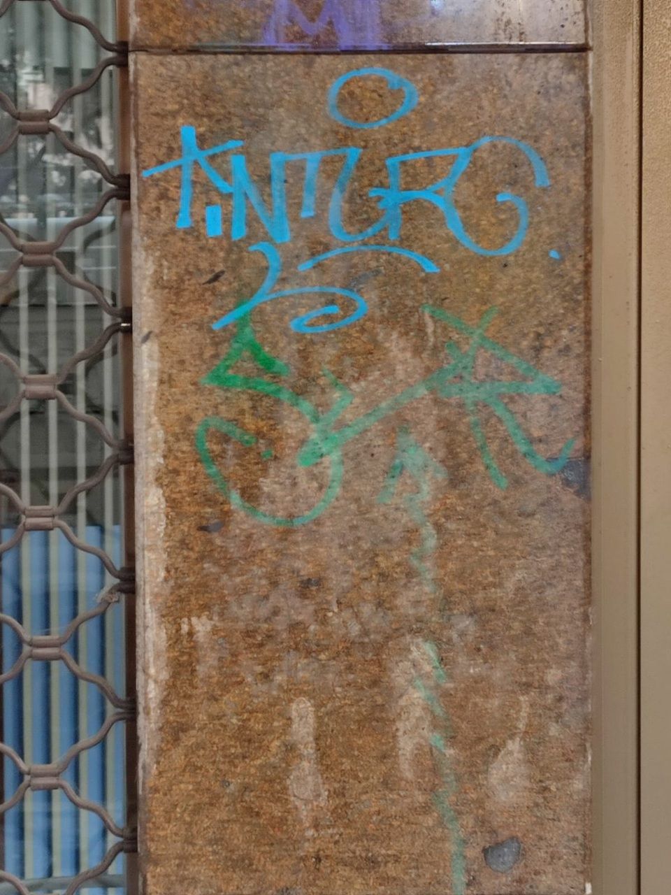 Pilastro di cemento consumato dagli agenti atmosferici con graffiti blu e verdi accanto a una recinzione a rete metallica.