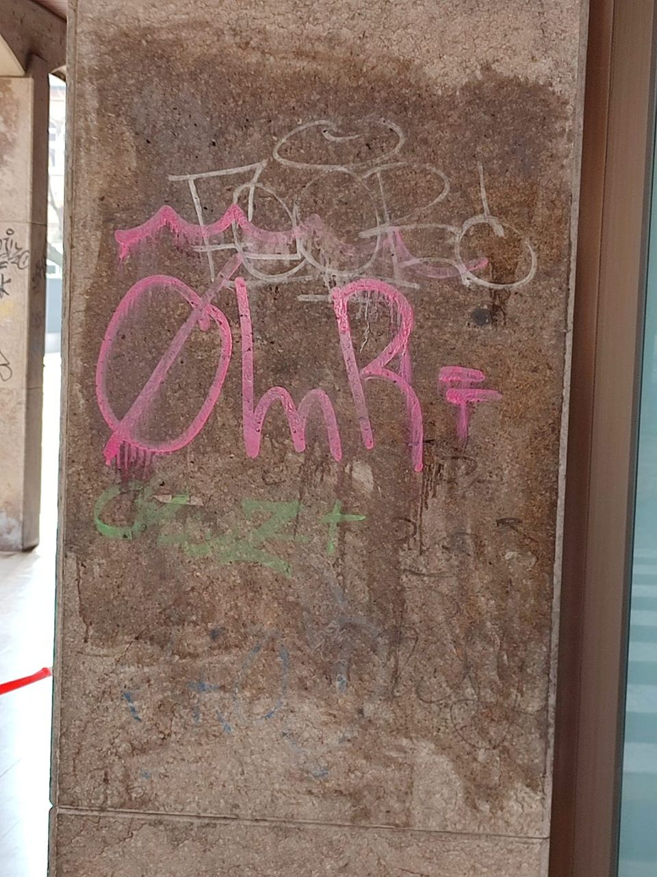 Muro di cemento marrone con graffiti rosa e scarabocchi bianchi, visto da uno stretto vicolo.