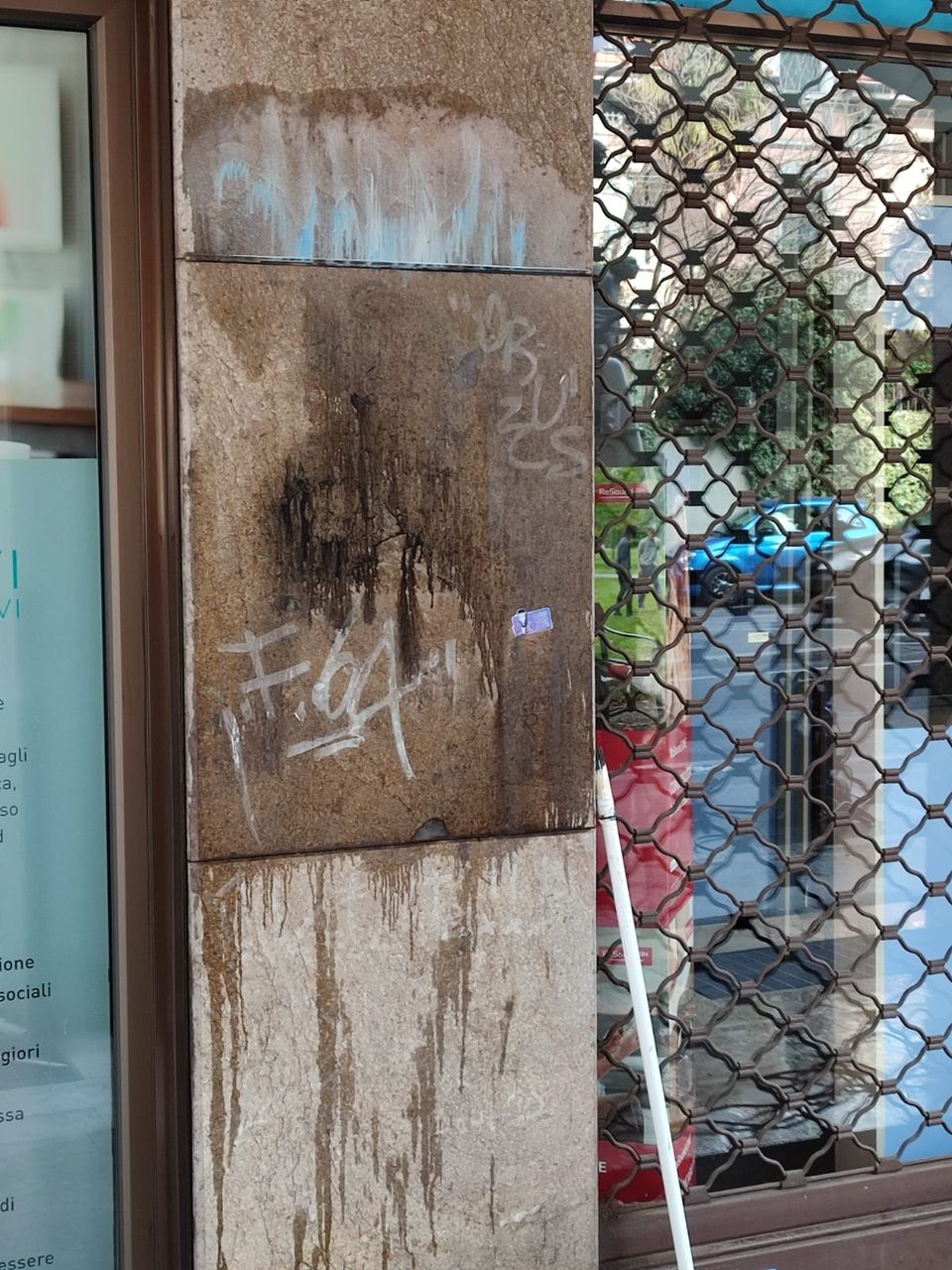 Un pilastro di pietra imbrattato di graffiti accanto a una porta a vetri e a una recinzione a rete metallica, con oggetti da esterno alle sue spalle.
