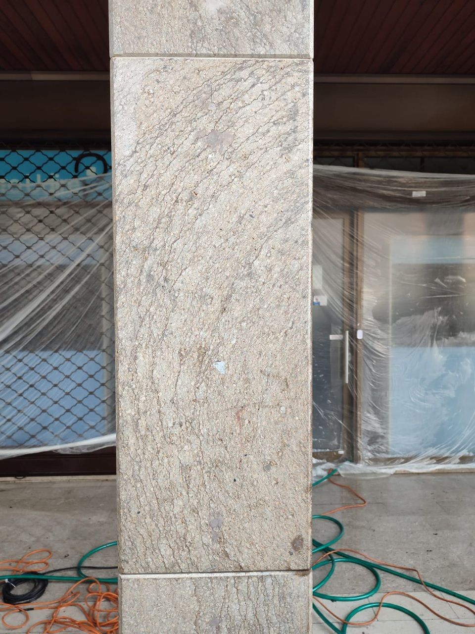 Sullo sfondo si vedono una colonna di pietra, una rete da cantiere, un telo di plastica e dei tubi flessibili arrotolati.