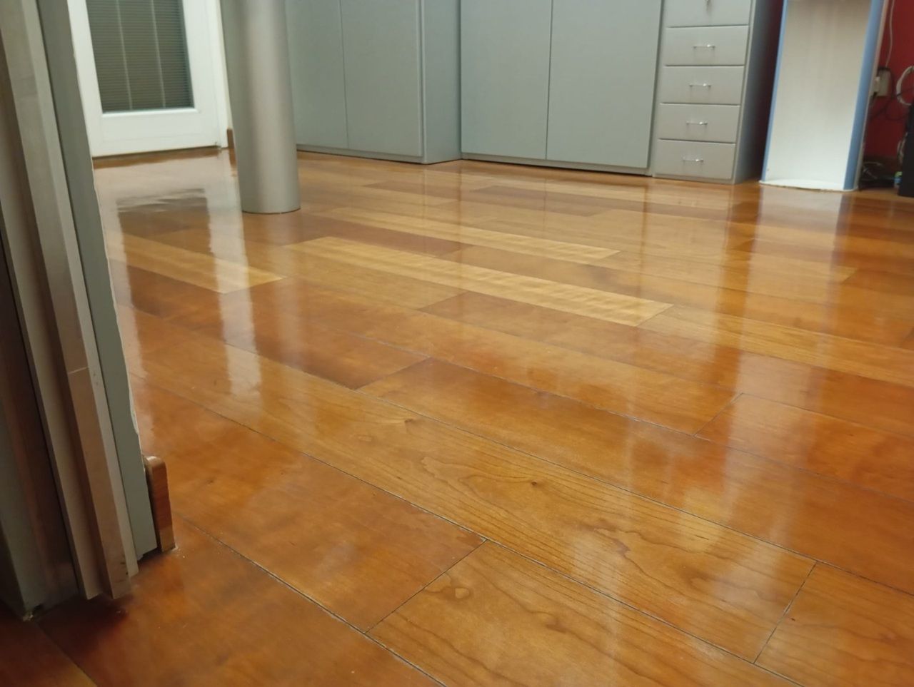 Pavimento in legno lucido nel corridoio, con riflessi che conducono a una porta e ad armadi sullo sfondo.
