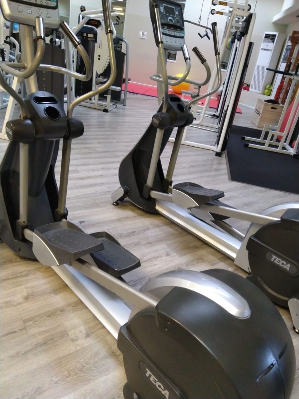 Due ellittiche in una palestra, con struttura argentata e rivestimento nero, su un pavimento in legno.