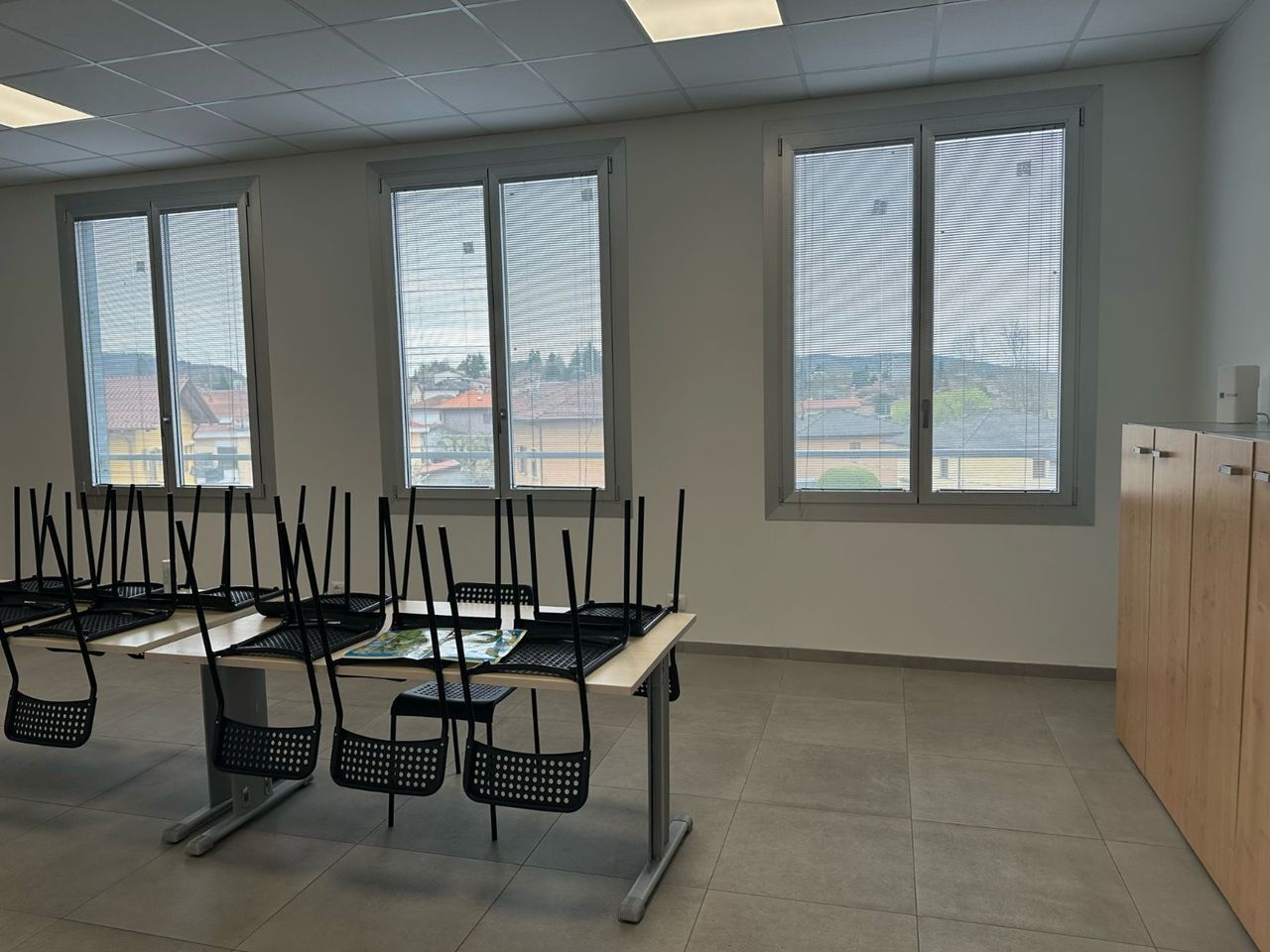 Aula vuota con banchi capovolti sui tavoli, grandi finestre e armadietti beige lungo la parete.