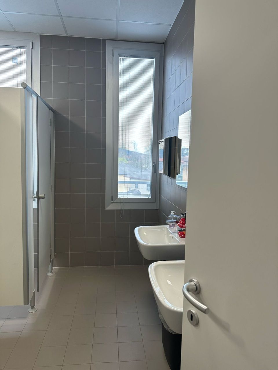 Piccolo bagno pubblico con pareti rivestite di piastrelle grigie, box doccia, lavandino e porta aperta.